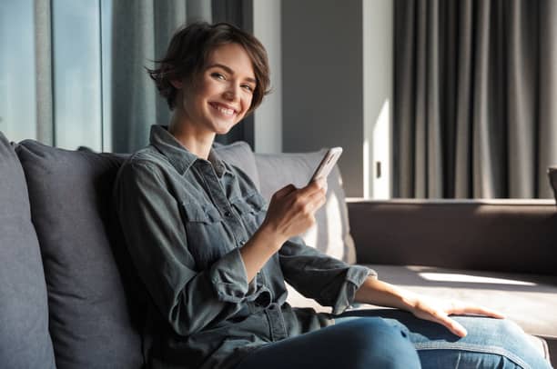 Hymyilevä nainen istuu sohvalla älypuhelin kädessään